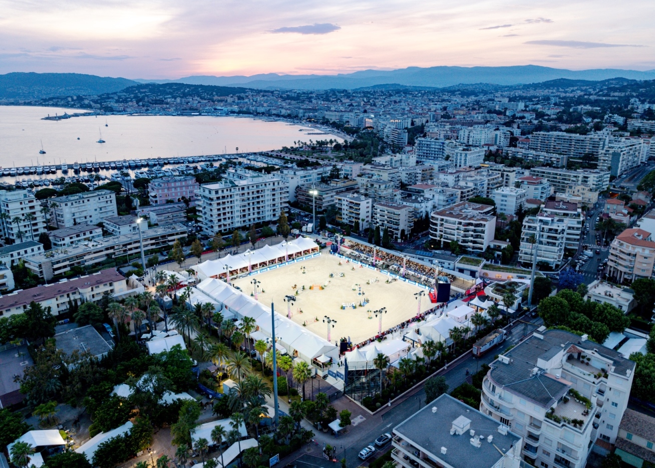 view of the Longines Global Champions Tour arena in Cannes, set against the French Riviera skyline with grandstands, floodlit jumps, and luxury yachts in the background near La Croisette