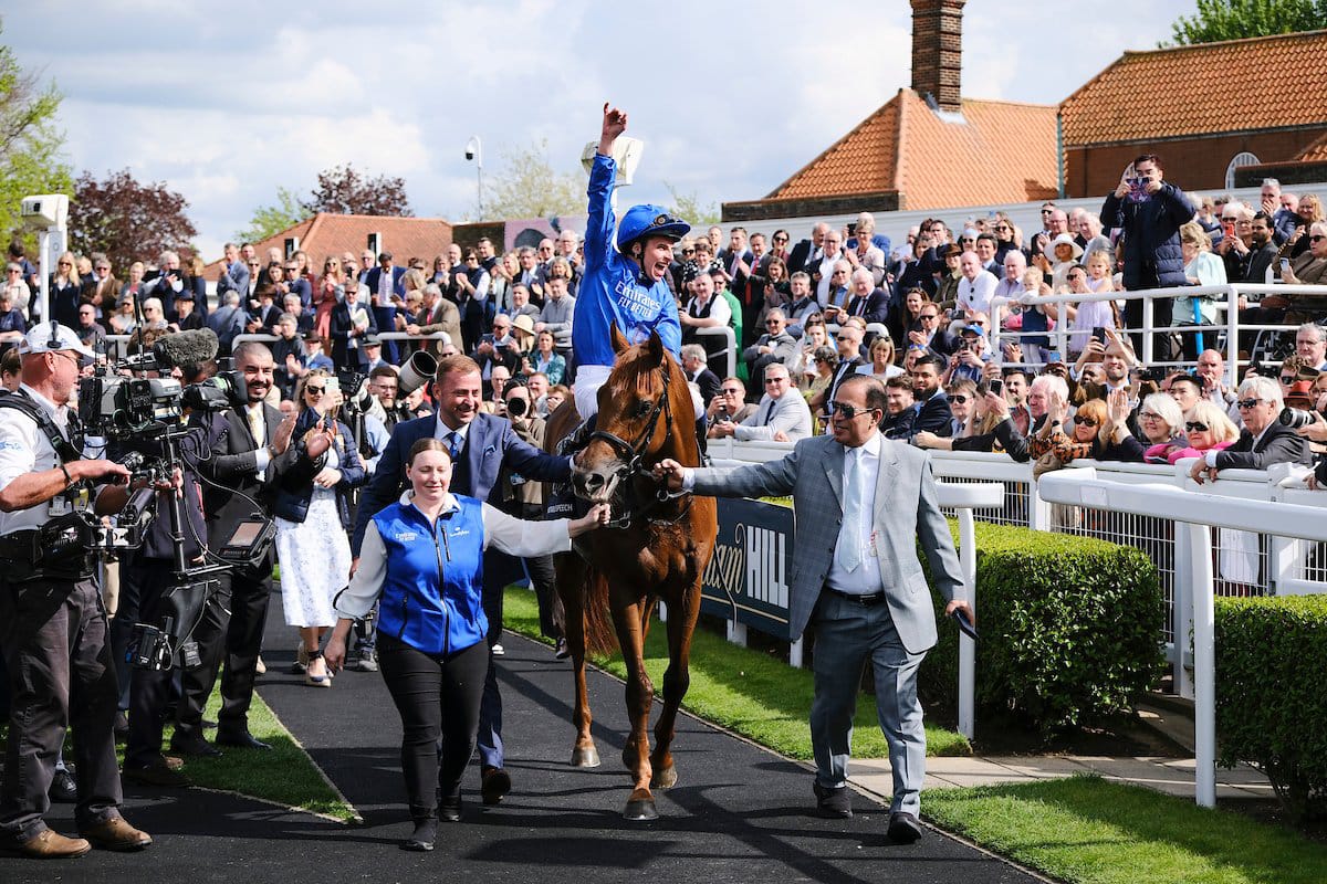 A panoramic view of Newmarket Racecourse with racehorses in action during the Guineas Festival, surrounded by cheering spectators under a bright spring sky.