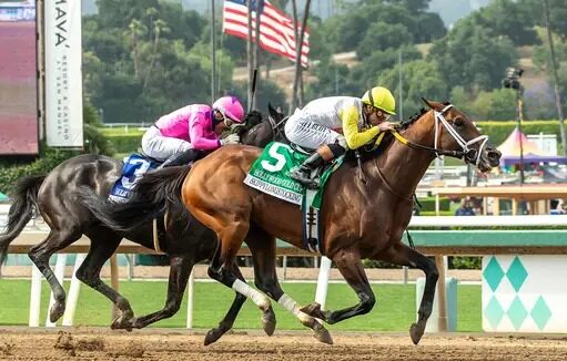 Aerial view of Santa Anita Park with horses racing down the track on a sunny day, showcasing a packed grandstand and scenic mountain backdrop during a major race day event on May 30, 2025.