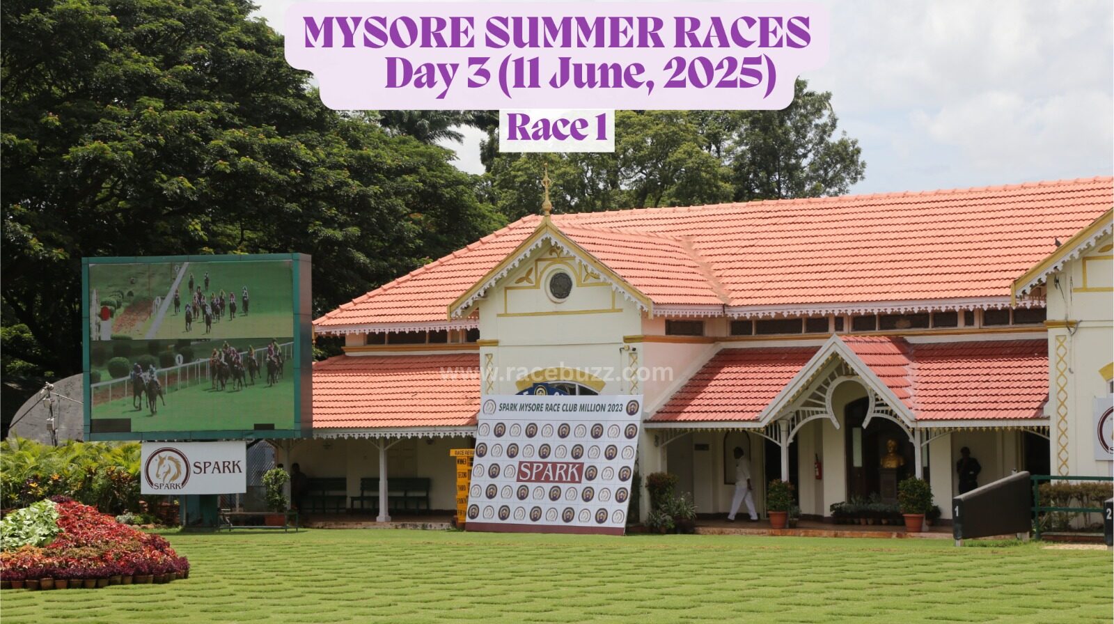 The image shows the Mysore Race Club paddock area during the Mysore Summer Races 2025 – Day 3 on June 11, before Race 1. A large digital screen on the left displays the live race feed, while the race club building with its traditional red-tiled roof and colonial architecture stands in the background. A sponsorship backdrop from Spark is placed prominently on the lawn, which is neatly maintained with decorative grass patterns.