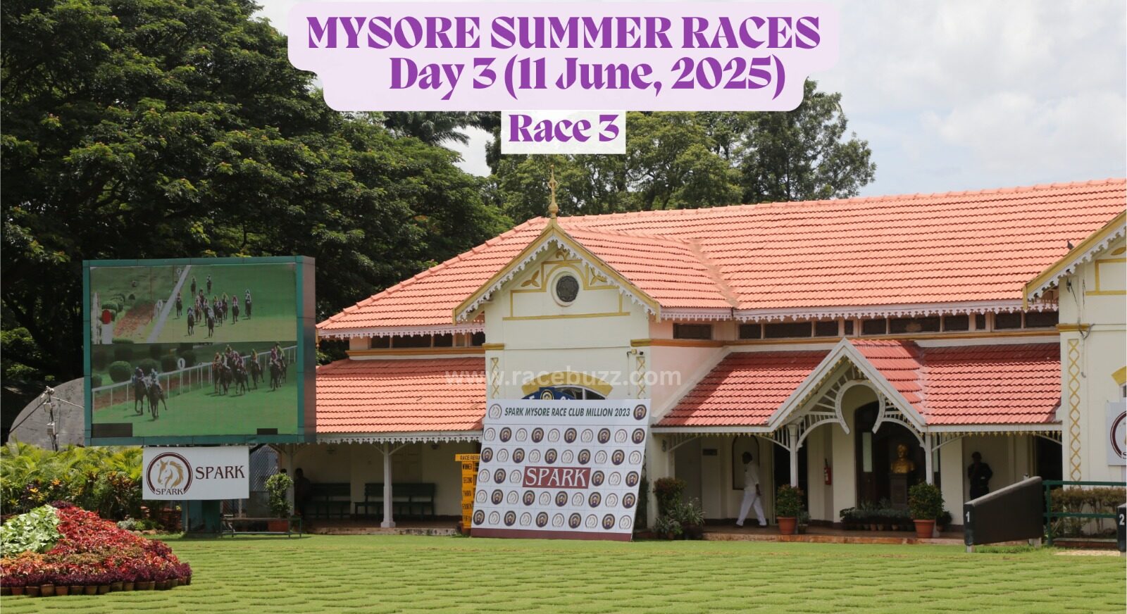 The image shows the Mysore Race Club paddock area during the Mysore Summer Races 2025 – Day 3 on June 11, before Race 3. A large digital screen on the left displays the live race feed, while the race club building with its traditional red-tiled roof and colonial architecture stands in the background. A sponsorship backdrop from Spark is placed prominently on the lawn, which is neatly maintained with decorative grass patterns.