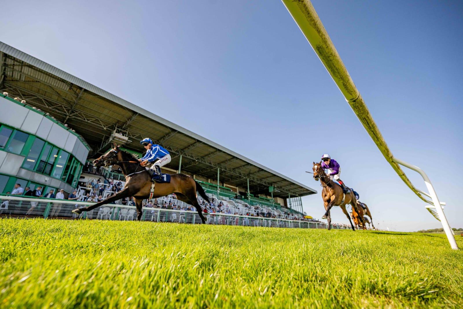 Horses racing at Brighton Racecourse during the Summer Afternoon Racing event with jockeys in colorful silks on a bright sunny day.